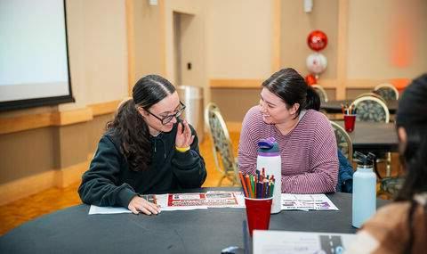 Two women sit at a table looking at and talking about materials on the table with BSU balloons in background